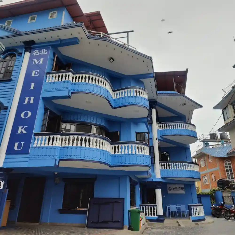 Blue, three-story building with ornate white railings and a sign reading "MEIHOKU" in white text. Overcast sky with a bird flying above.