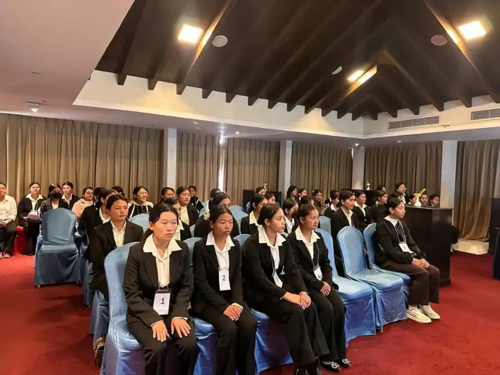A group of people in formal black suits with white shirts sit attentively in rows on blue chairs in a conference room. The scene is focused and professional.