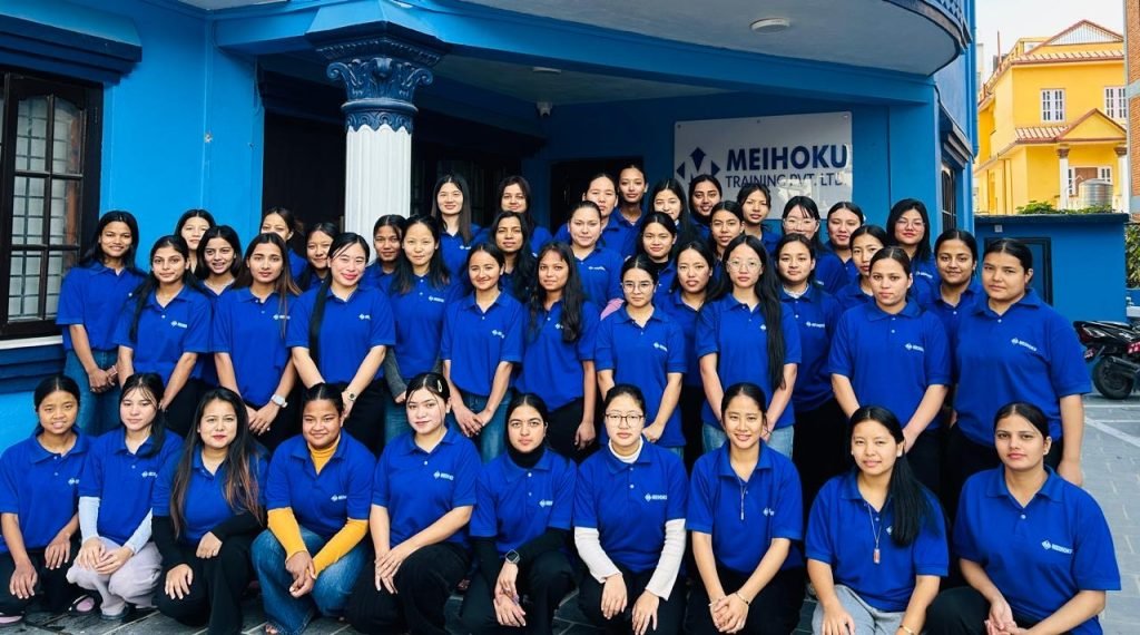 A group of thirty-six women in blue shirts stand together, smiling, in front of a blue building with "Meihoku Training" signage. The mood is positive and unified.