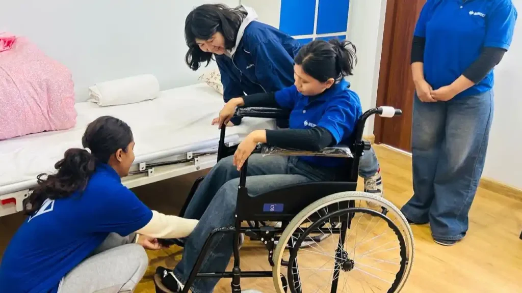 Three women assist a person in a wheelchair next to a bed. They all wear blue shirts, working together with care and focus in a bright room.