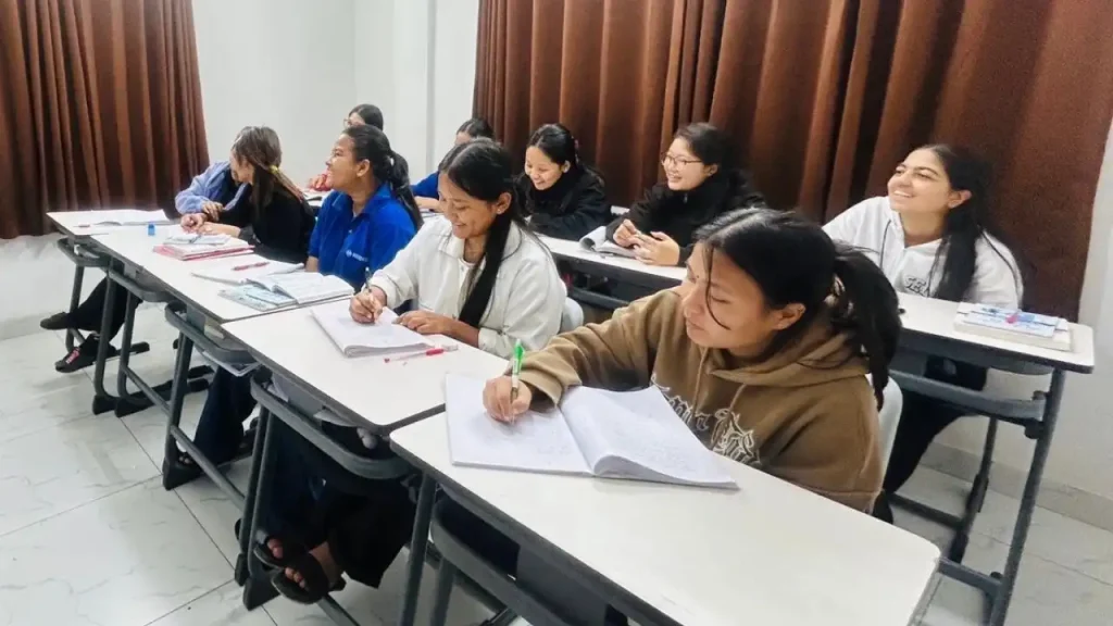 A group of eight students are seated at desks in a classroom, eagerly taking notes and smiling. The room has white walls and brown curtains, creating a warm, focused atmosphere.