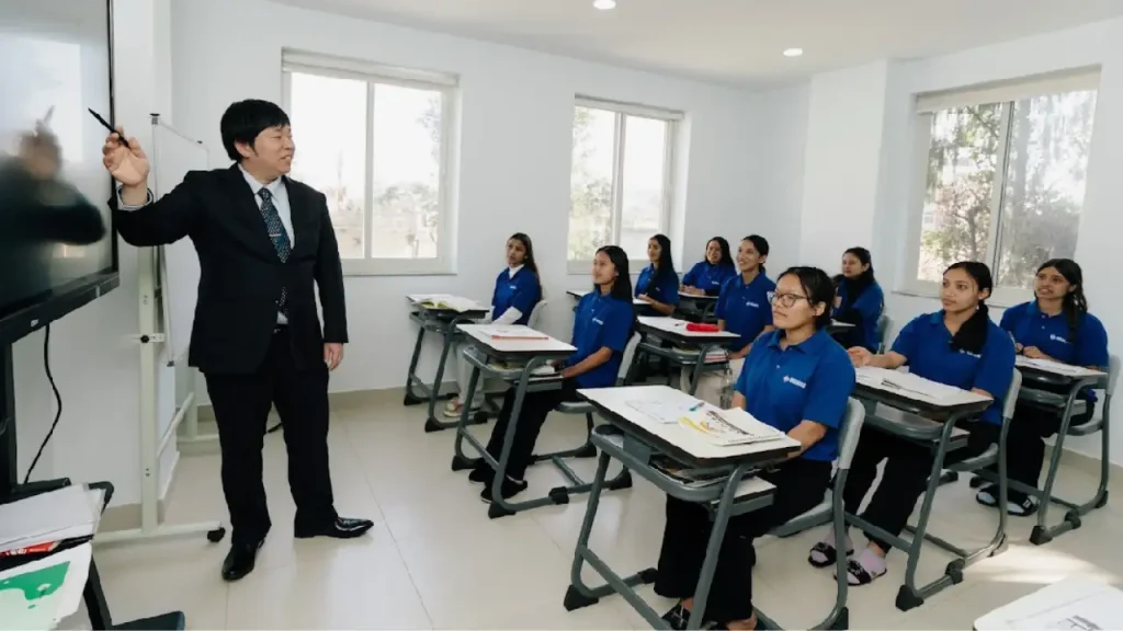 A teacher in a suit points at a screen in a bright classroom. Nine students in blue uniforms sit at desks, attentively listening. The mood is focused.