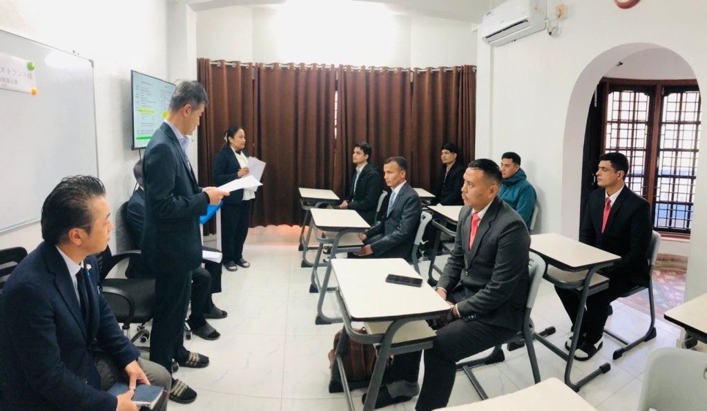 A group of men in suits sit at desks in a classroom while a man and woman stand at the front, presenting. The room has a focused and formal atmosphere.