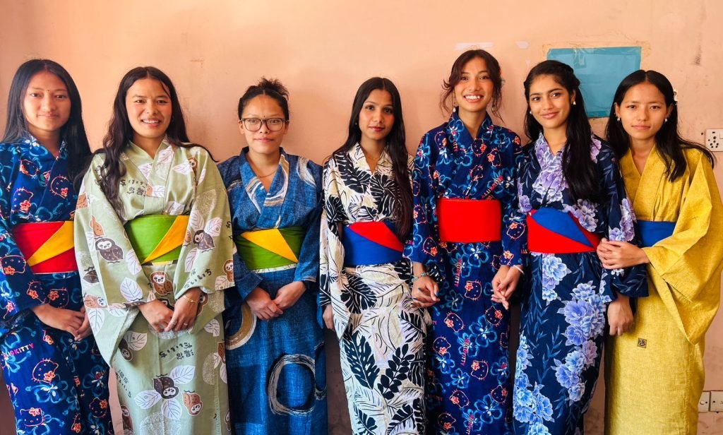 Seven women stand side by side, smiling, wearing vibrant kimonos with colorful obi sashes. The mood is joyful and celebratory.