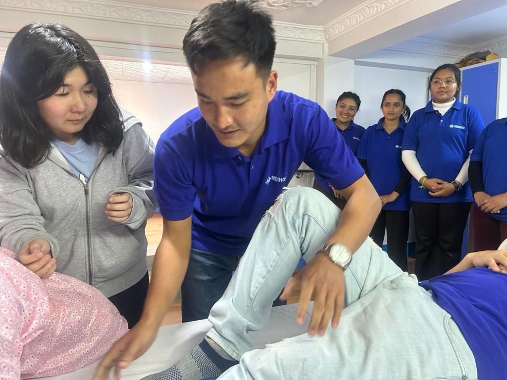 A group of people in blue shirts observe as a man gently examines a person's knee on a table. The scene conveys a teaching atmosphere.