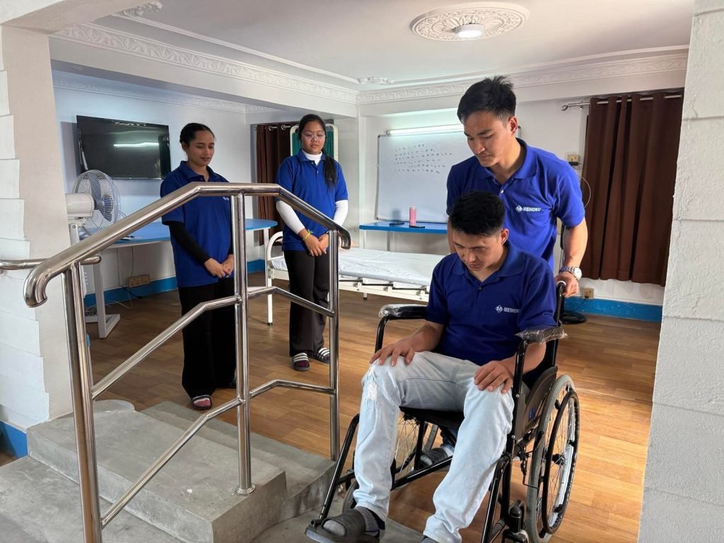 A caregiver assists a person in a wheelchair near stairs, while two other caregivers observe. The room is bright with wooden flooring and a whiteboard.