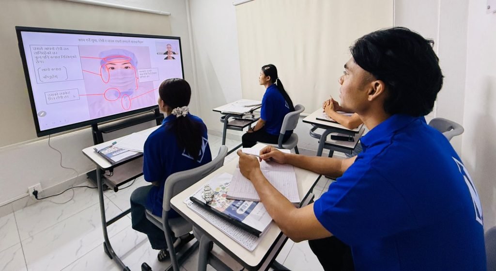 Classroom setting showing students in blue uniforms seated at desks, focused on an interactive digital display screen presenting a lesson in Nepali on proper workplace safety attire, including a cap and face mask.