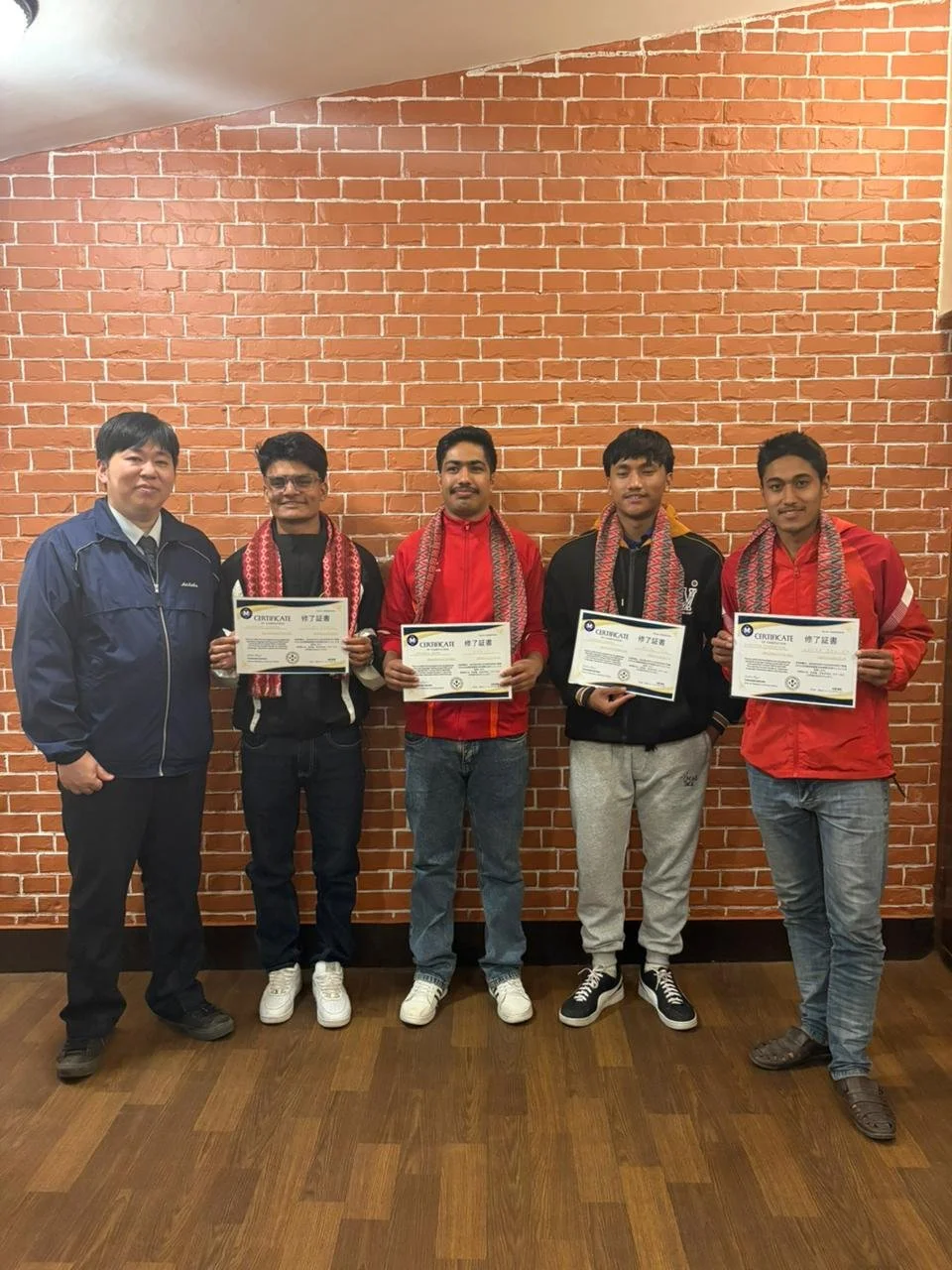 Four Nepali students wearing traditional Dhaka scarfs stand in front of a brick wall proudly holding completion certificates alongside their instructor.
