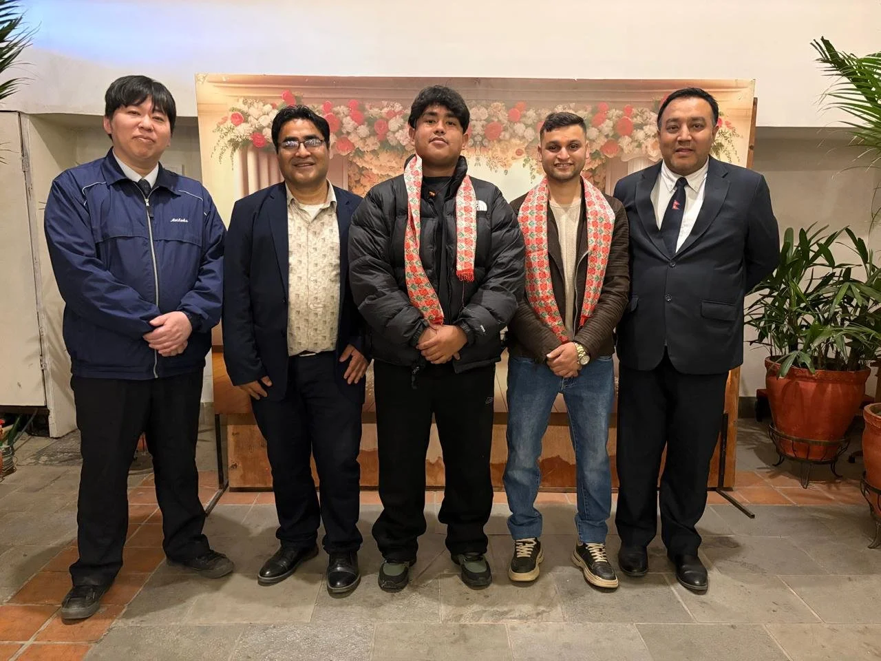 Group photo featuring two students wearing traditional Nepali Dhaka scarfs standing alongside three instructors and officials in an indoor setting.